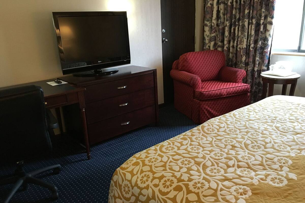 Une chambre d’hôtel avec un lit, un bureau avec une télévision, un fauteuil rouge et une petite table.