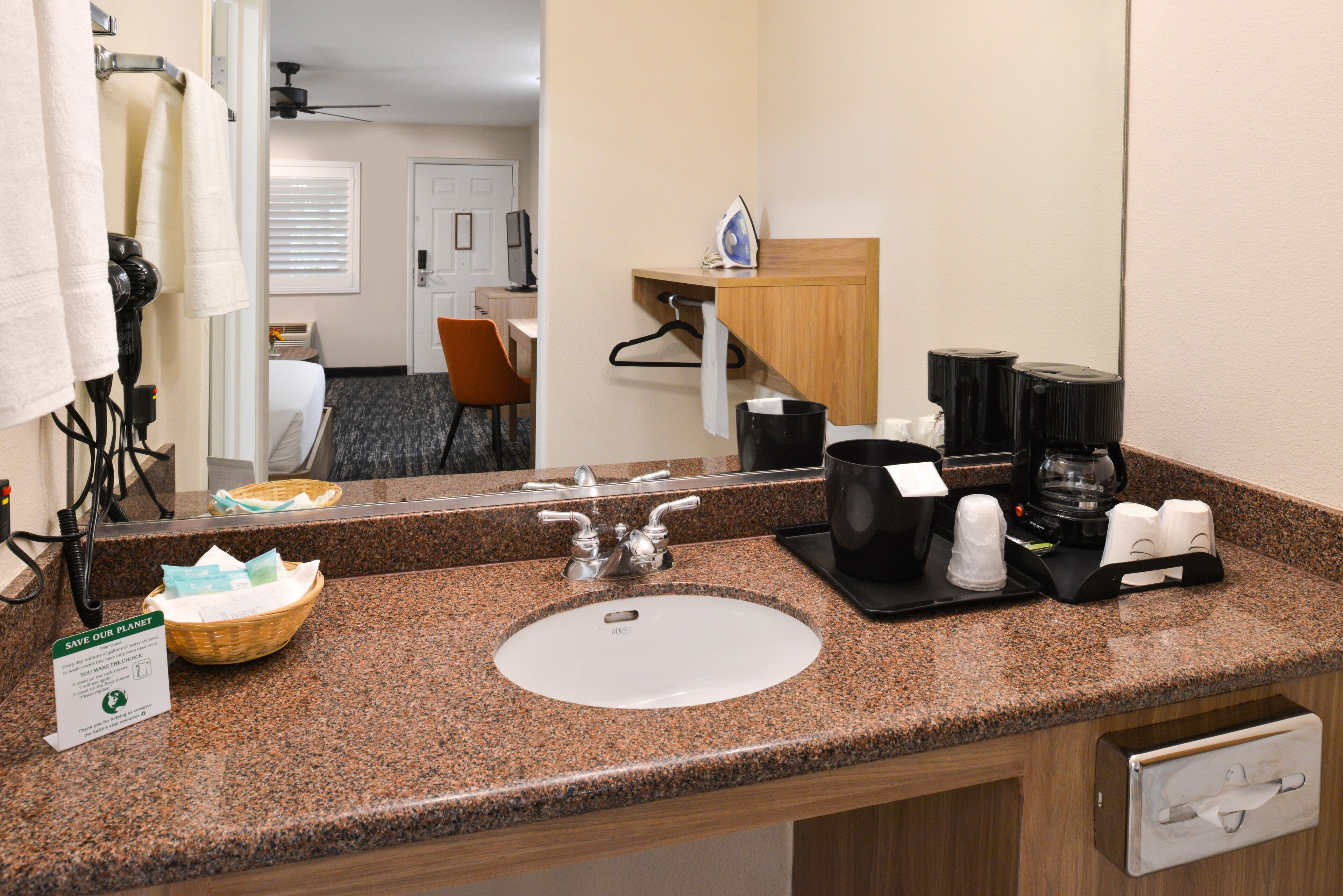 A hotel bathroom with a granite countertop, sink, and mirror. There is a towel rack, a basket, and a sign about saving the planet.