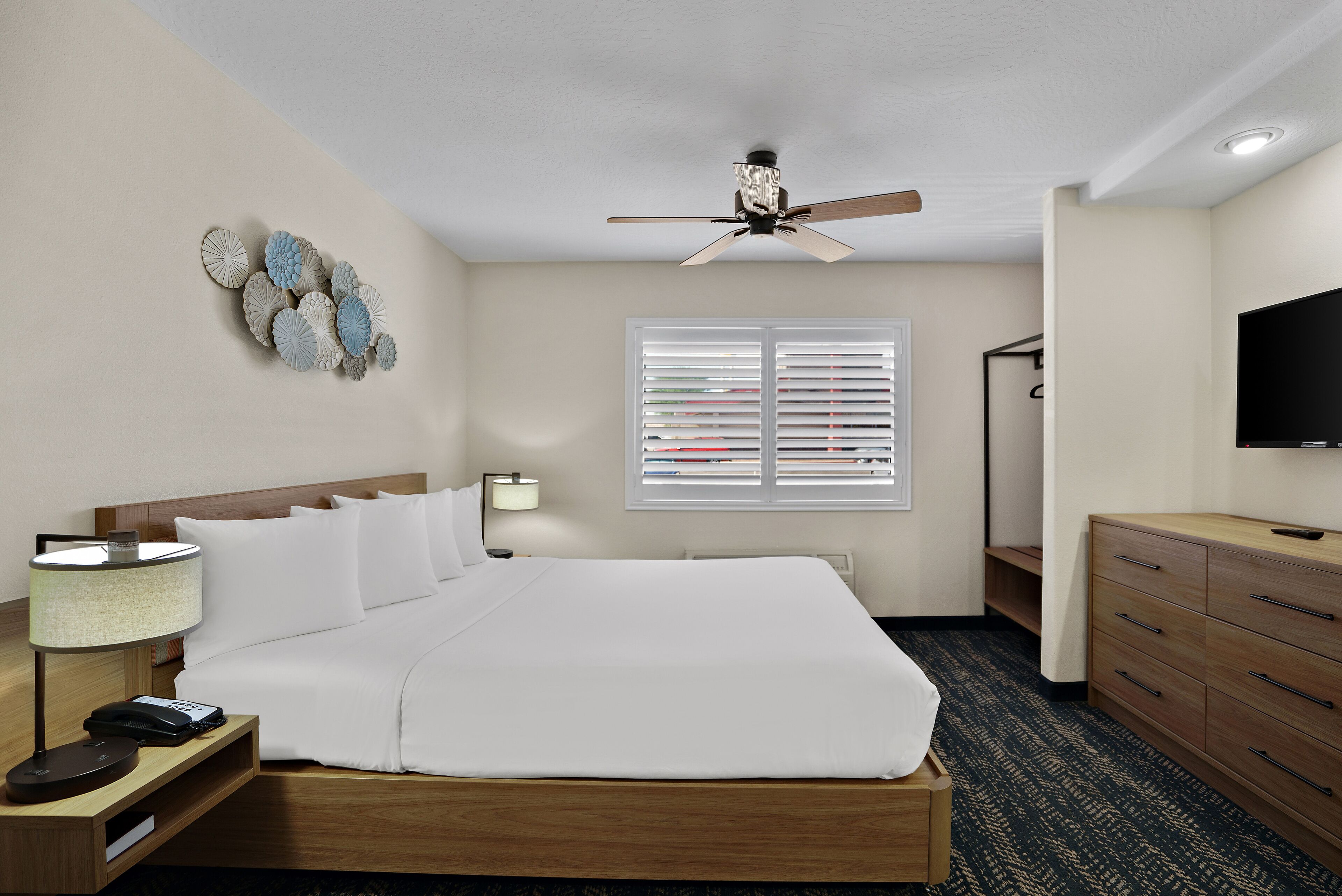 Habitación de hotel con cama, mesita de noche, lámpara, ventilador de techo, ventana con persianas, cómoda y televisor.