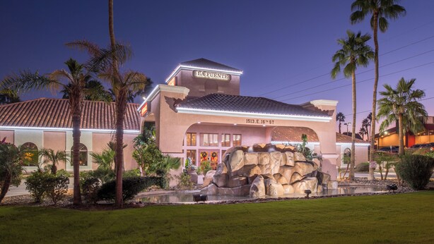 A hotel with a prominent sign reading "LA FUENTE," featuring a large rock waterfall in the front courtyard, surrounded by palm trees and well-maintained landscaping. - La Fuente Inn & Suites (Yuma)