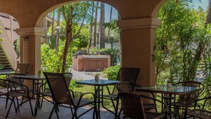 A covered outdoor seating area with tables and chairs, surrounded by greenery and a view of a building. - La Fuente Inn & Suites (Yuma)
