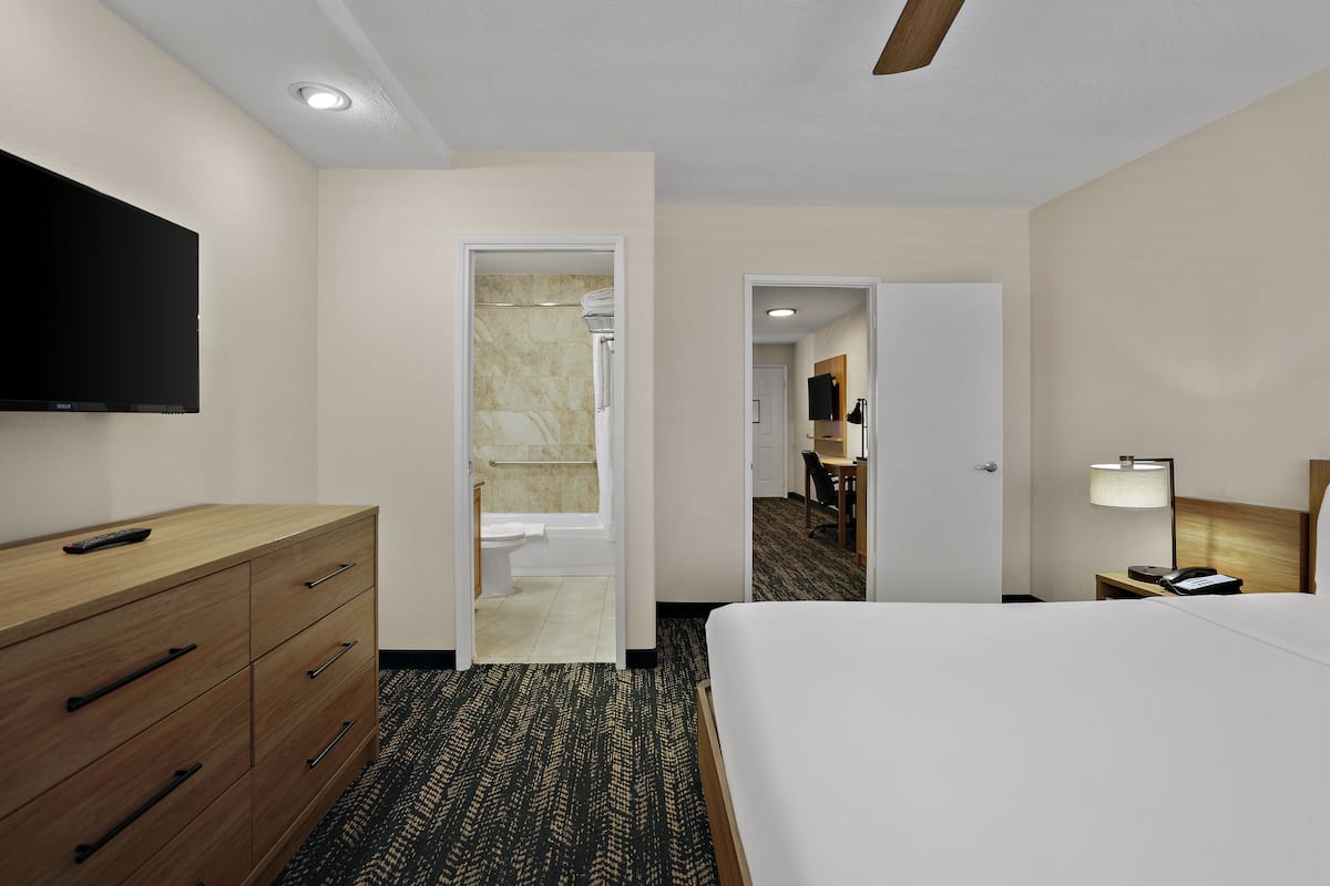 Une chambre d’hôtel comprenant un lit, une commode, une télévision, une salle de bain et un bureau avec une lampe.
