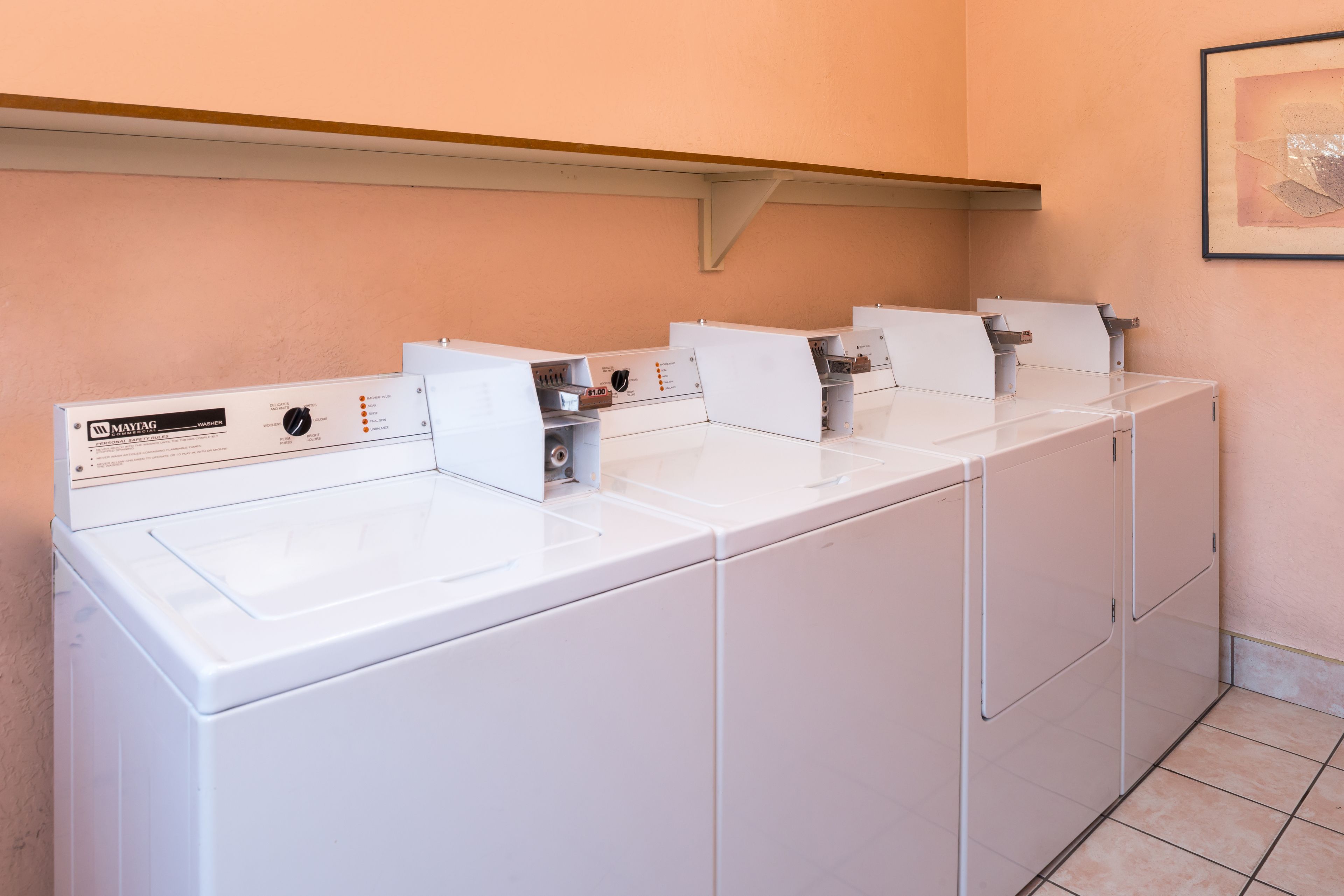 Une rangée de lave-linge Maytag à chargement frontal, de couleur blanche, dans une buanderie avec un mur de couleur pêche et un tableau encadré.