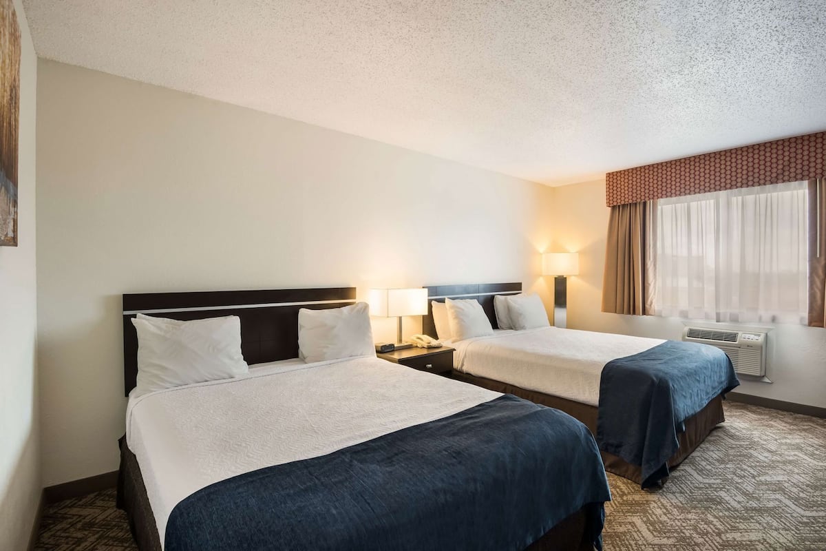 A hotel room with two beds, a nightstand with a lamp, a window with curtains, and an air conditioning unit.