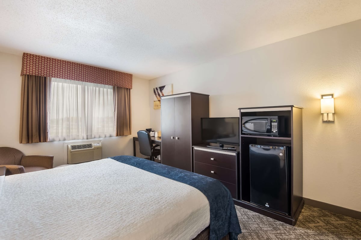 A hotel room with a bed, a desk with a computer, a microwave, a refrigerator, and a chair.