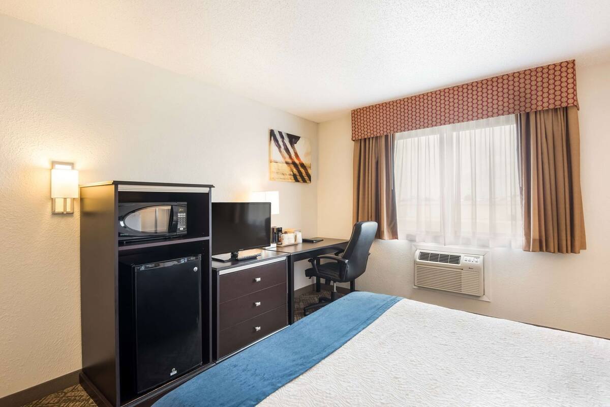 A hotel room with a bed, desk, chair, microwave, television, and a window with curtains.