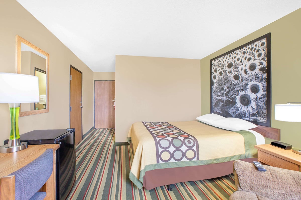 A hotel room with a bed, a desk with a lamp, a chair, a large mirror, and a wall with a sunflower painting.