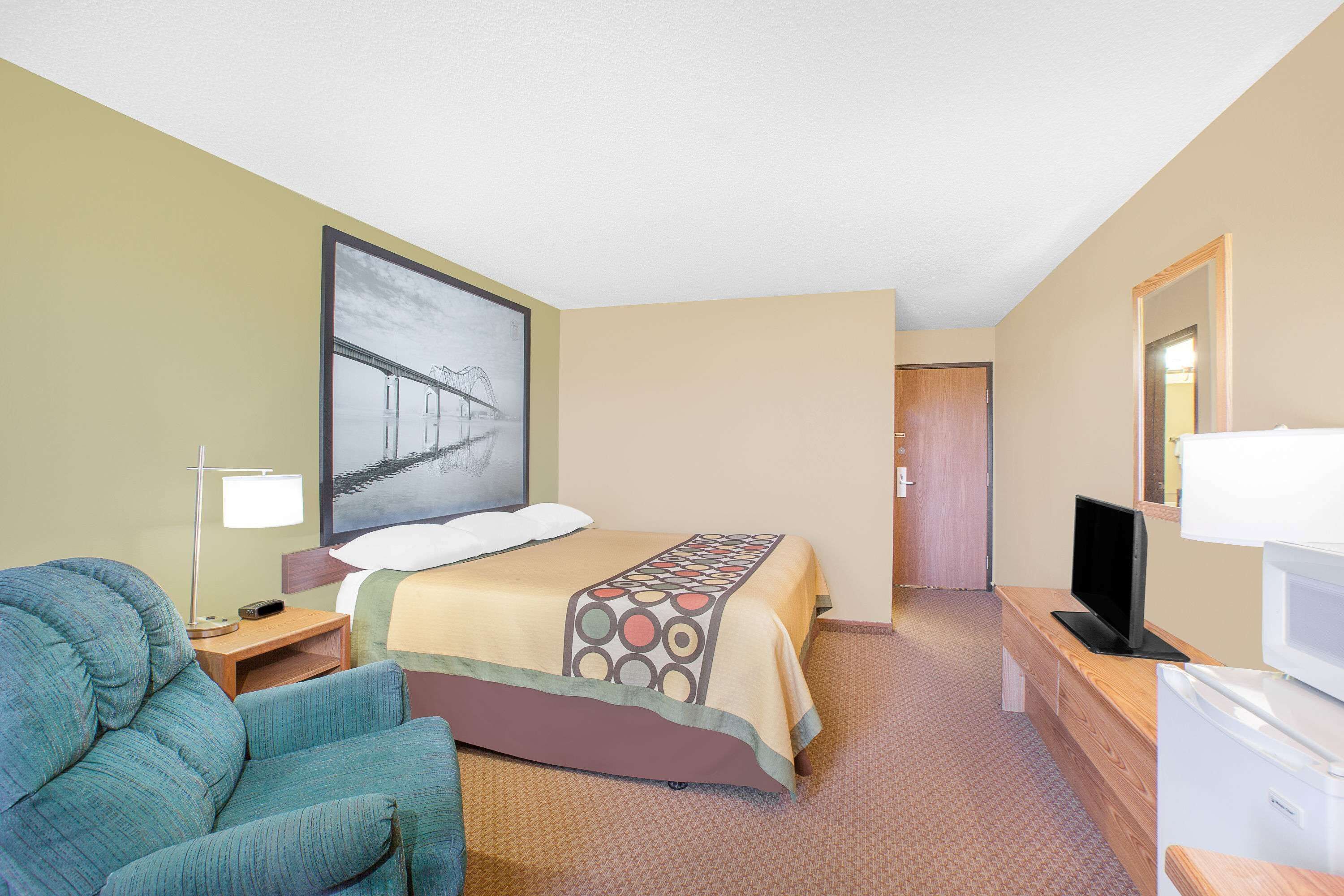 Quarto de hotel com cama, televisão, escrivaninha, cadeira e um quadro na parede.