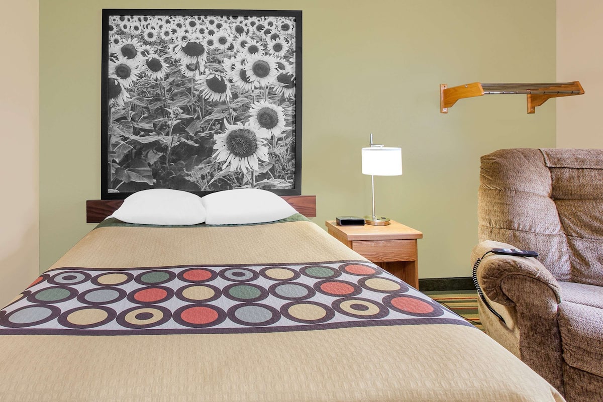 A neatly made bed with a colorful circular pattern, a framed black and white sunflower painting on the wall, a bedside lamp, and a brown armchair.