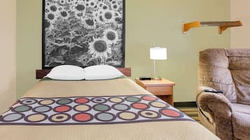 A neatly made bed with a colorful circular pattern, a framed black and white sunflower painting on the wall, a bedside lamp, and a brown armchair.