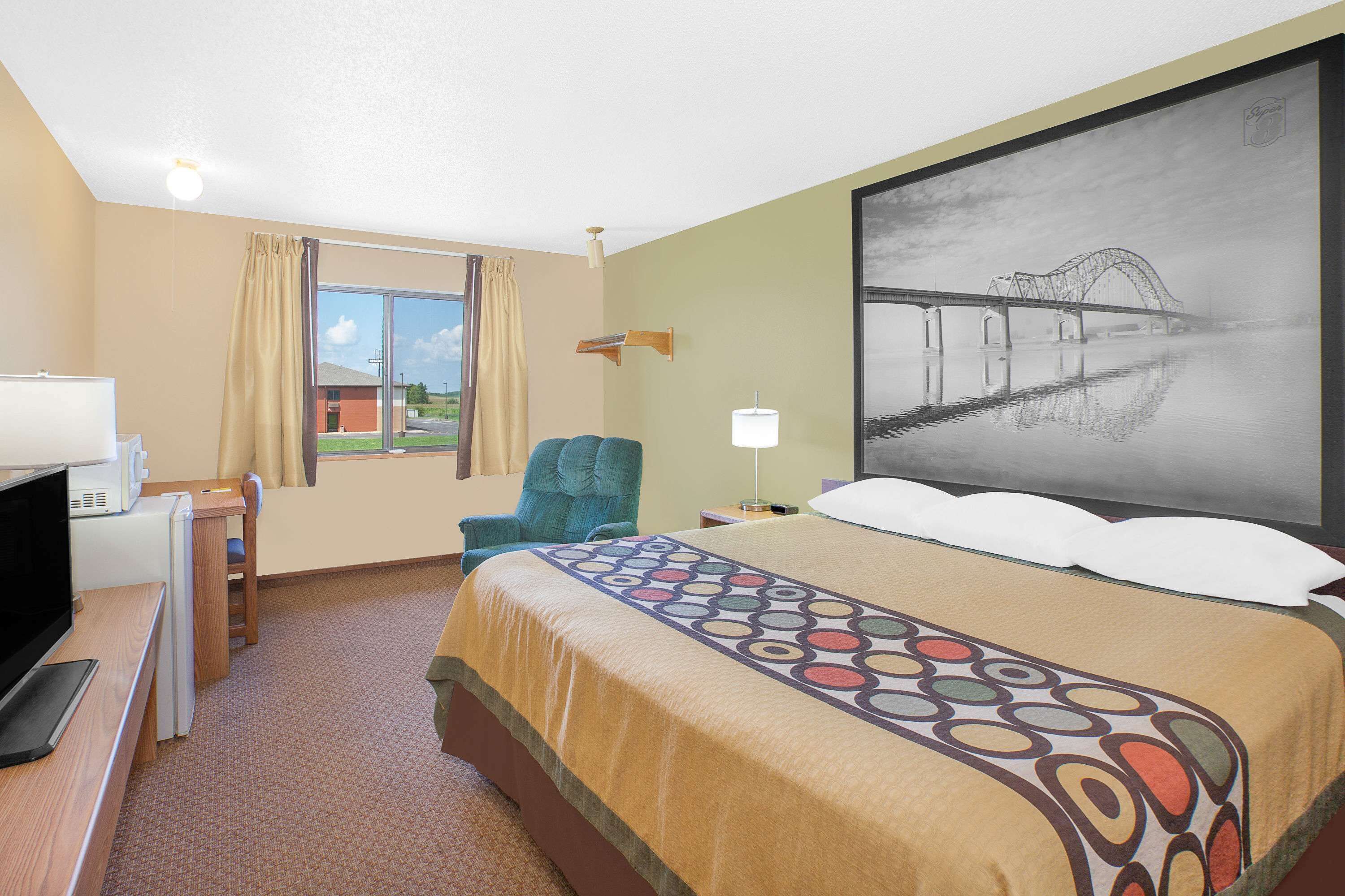 Une chambre d’hôtel avec un lit, un bureau avec un ordinateur, une chaise, une fenêtre avec des rideaux et un tableau représentant un pont.