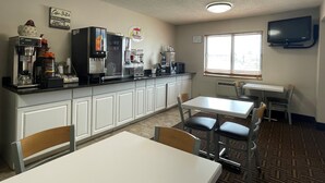 Una habitación pequeña y limpia con kitchenette, cafetera, microondas y televisor montado en la pared. Hay dos mesas con sillas y una ventana que deja entrar luz natural.