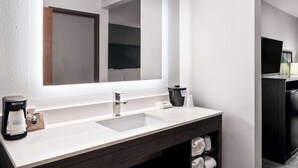 Un baño moderno con un espejo grande, un lavamanos blanco, una cafetera y un mueble de tonos oscuros.