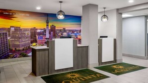 Zona de recepción de un hotel con un mural de la ciudad, dos mostradores de recepción y un felpudo verde que dice 'QUINTA'.