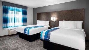 Habitación de hotel con dos camas, un ventanal grande con cortinas azules, una lámpara fijada a la pared y un cabecero moderno.