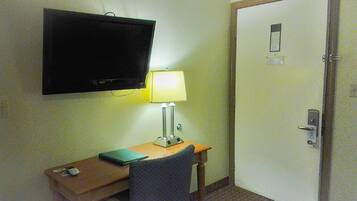 Et hotelværelse med et fjernsyn ophængt på væggen, et skrivebord med en lampe, en stol og en dør med lås.