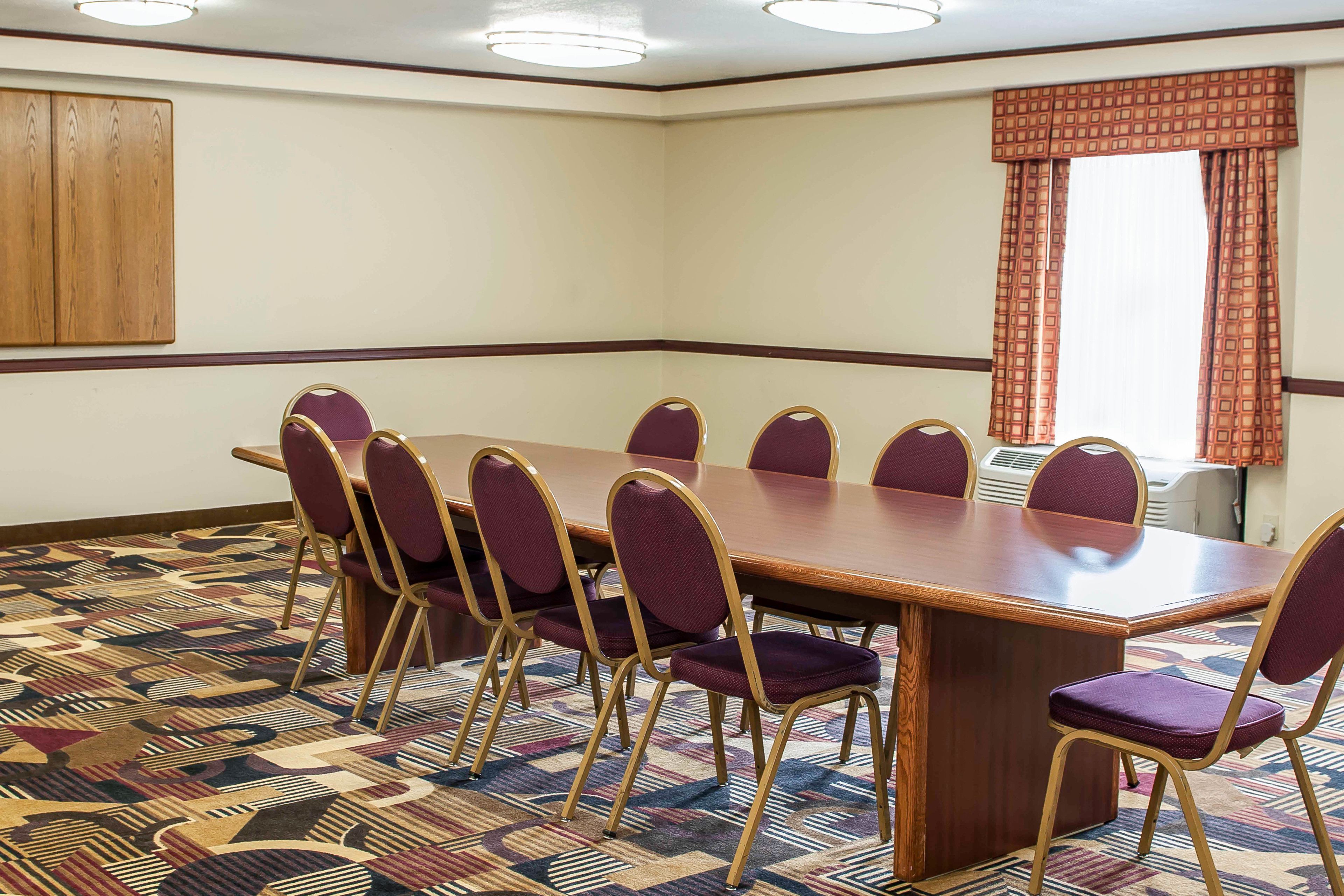 Une salle de conférence avec une longue table, huit chaises, un tapis à motifs, des panneaux de bois et une fenêtre avec des rideaux.