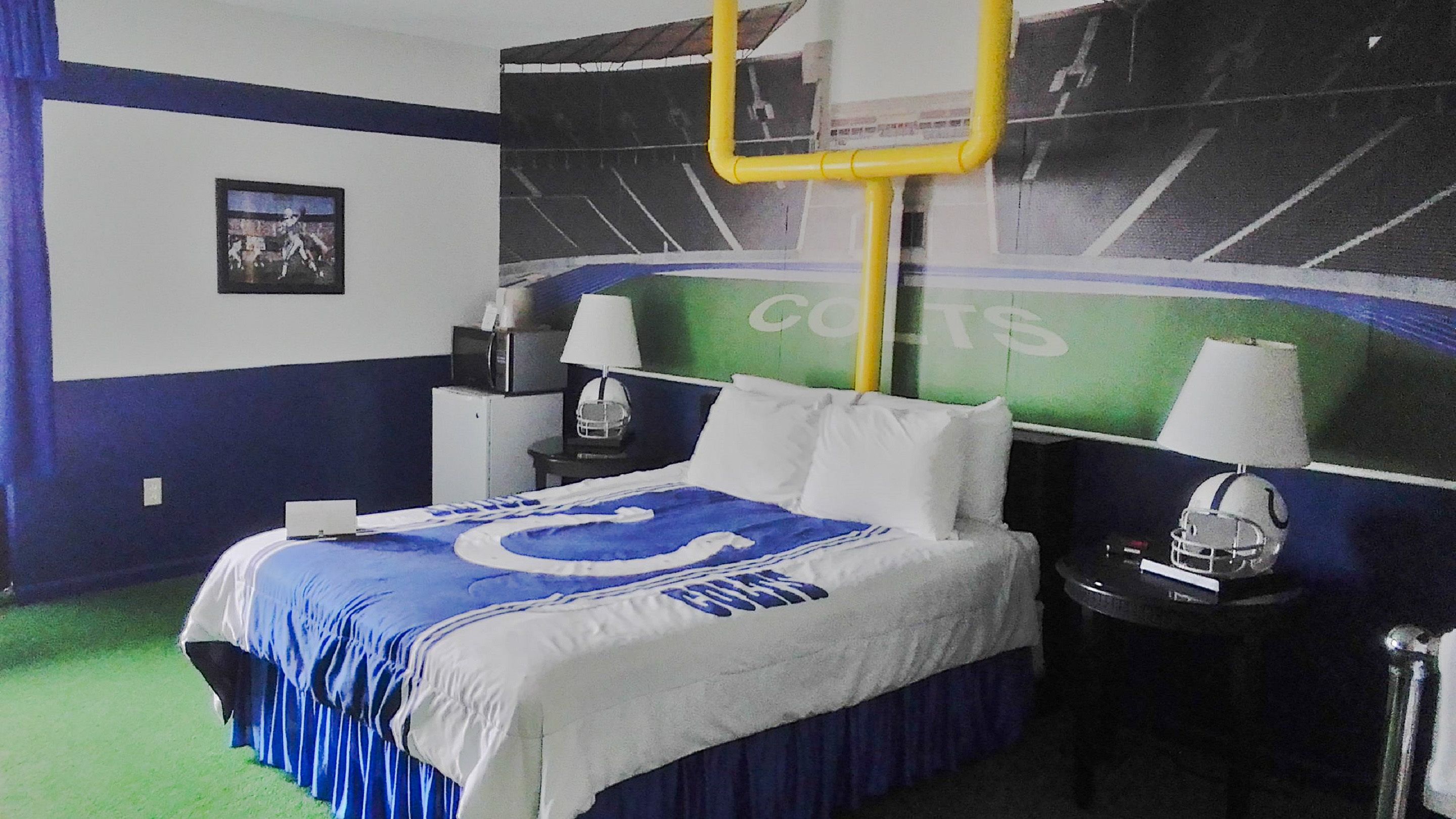Habitación de hotel con cama, dos mesitas de noche, lámpara, casco y un mural temático de deportes.