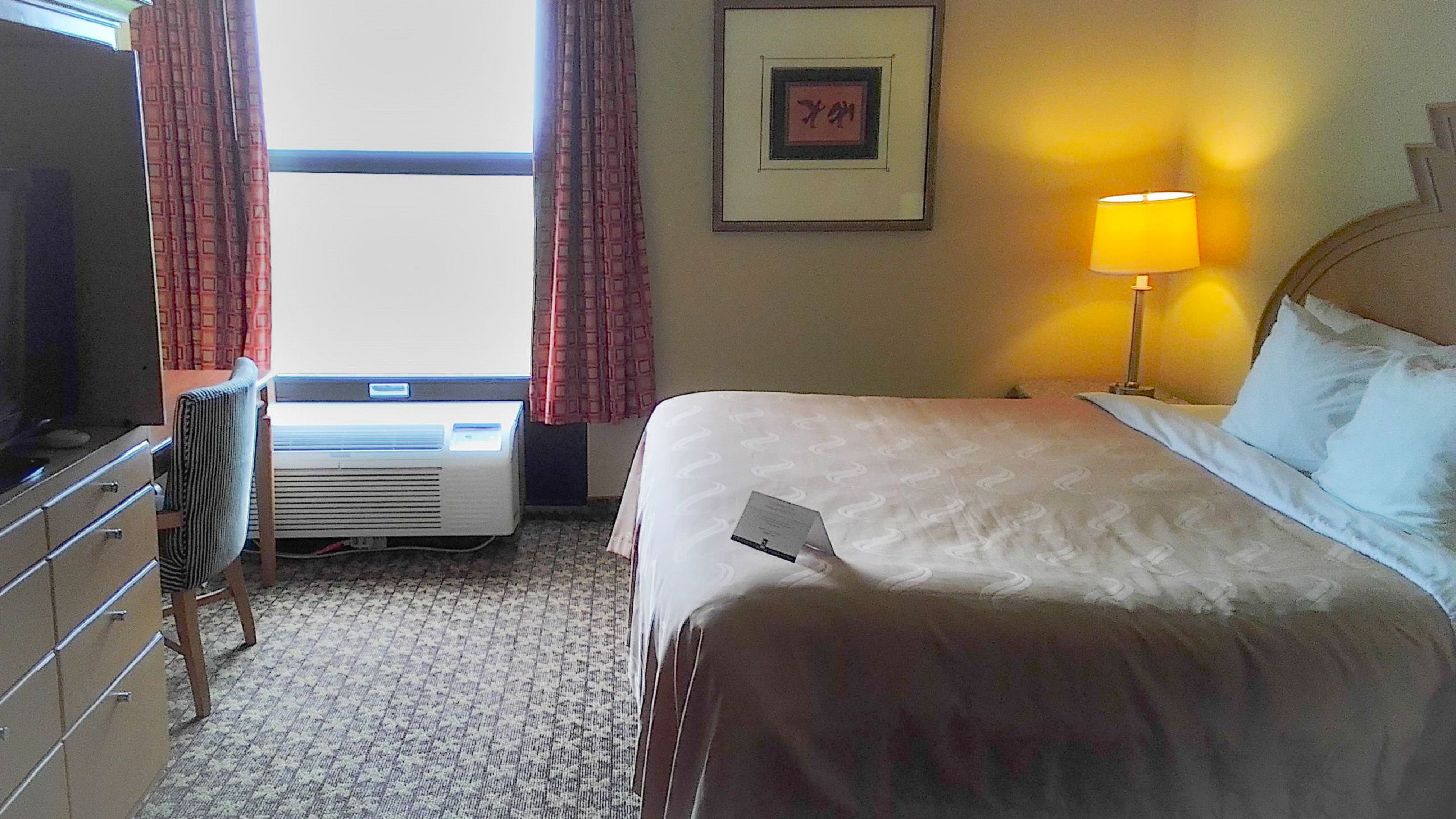 A hotel room with a bed, a desk, an air conditioner, a window with curtains, a framed picture on the wall, and a bedside lamp.