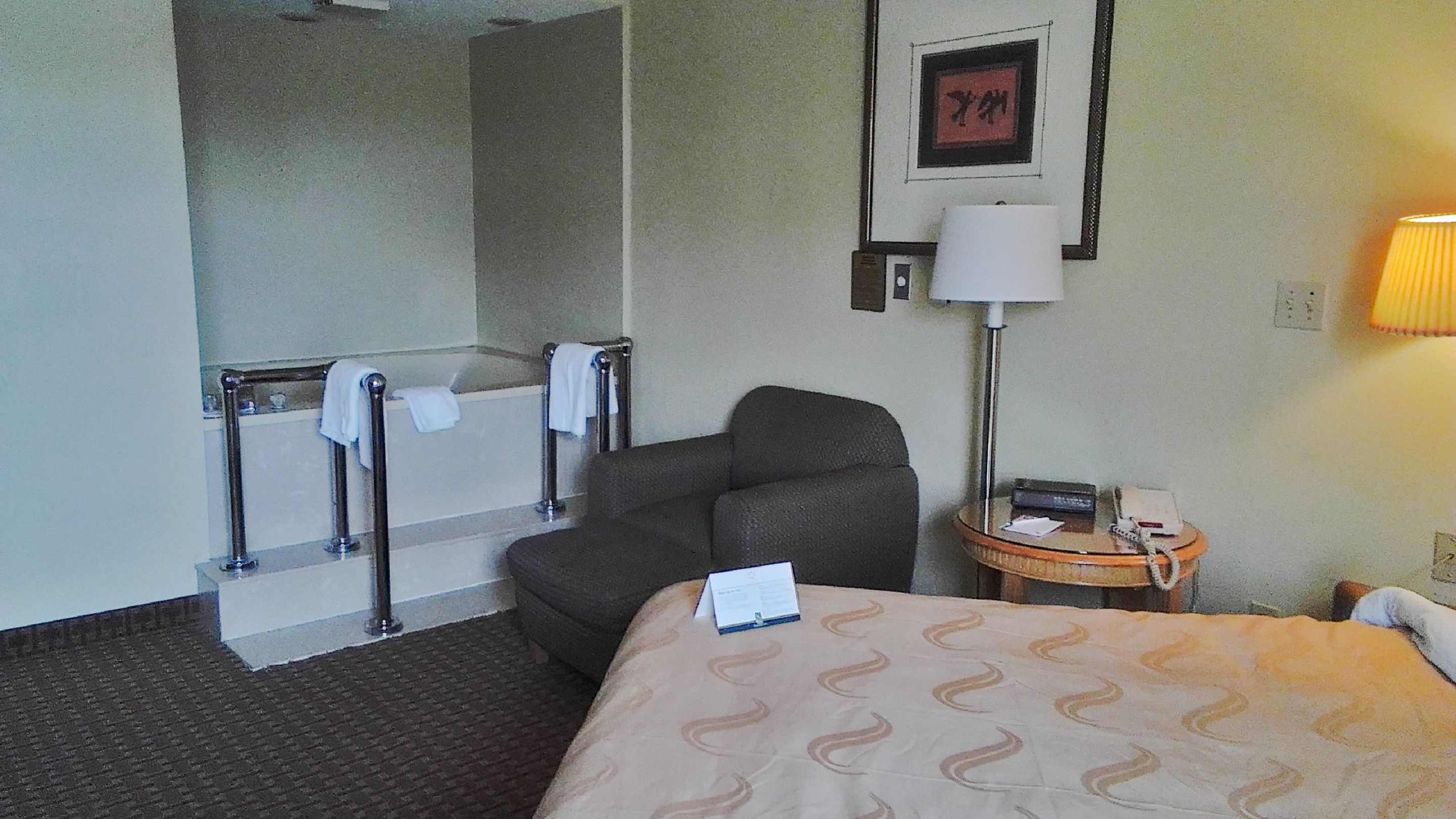 A hotel room with a bed, a chair, a small table with a lamp, a bathroom with a bathtub, and a towel rack.