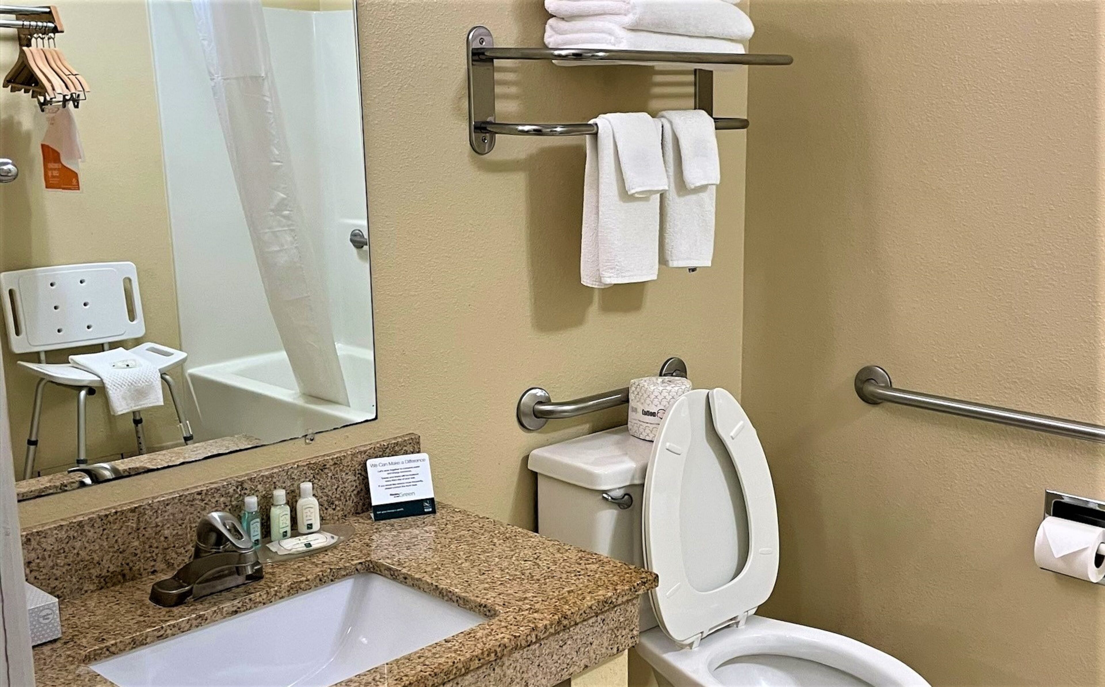 A bathroom with a sink, toilet, and handrails. There is a mirror, a towel rack, and a chair.