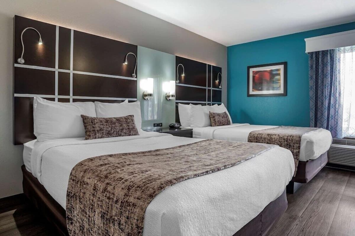 A hotel room with two beds, a large mirror, wall-mounted lights, a painting, and an air conditioning unit.