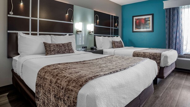 A hotel room with two beds, a large mirror, wall-mounted lights, a framed picture, and a window with curtains. - Best Western Carlton Suites (Birmingham)