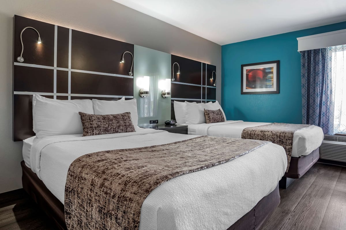 A hotel room with two beds, a large mirror, wall-mounted lights, a framed picture, and a window with curtains.