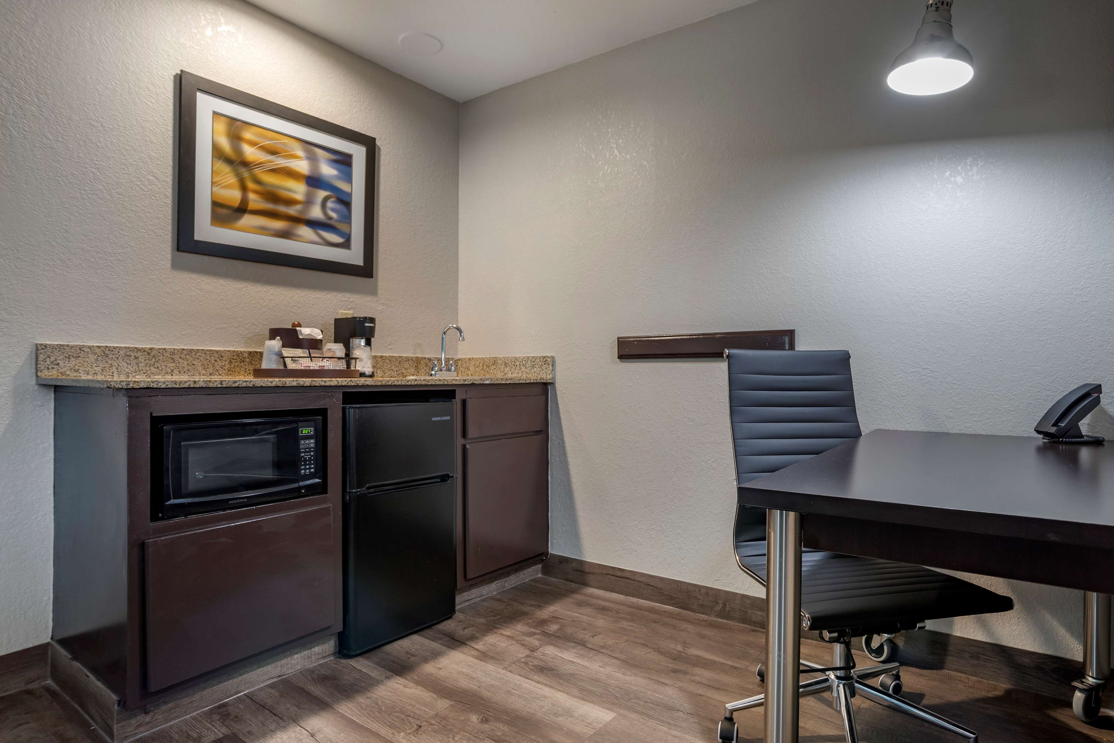 A compact kitchen with a microwave, refrigerator, and a coffee maker on the counter. There's a dining table with a chair and a wall-mounted shelf.