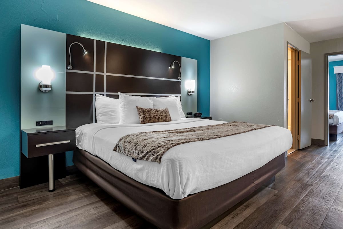 A modern hotel room with a large bed, a nightstand, a wall-mounted light, and a door leading to another room.