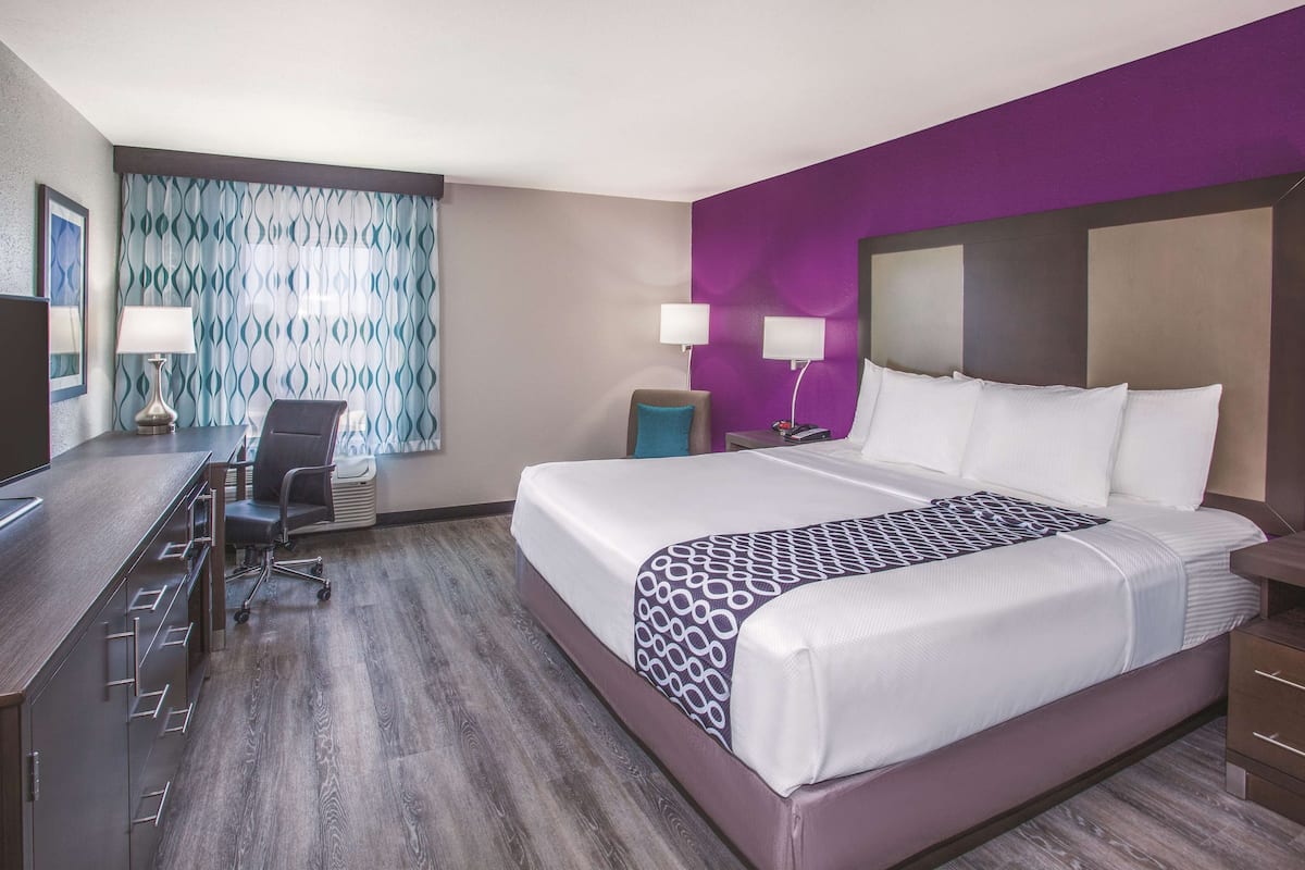 A hotel room with a large bed, a desk with a chair, two bedside lamps, and a window with patterned curtains.