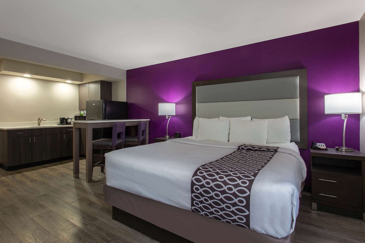 A hotel room with a large bed, a desk, a chair, a small table, a refrigerator, and a nightstand with a lamp.