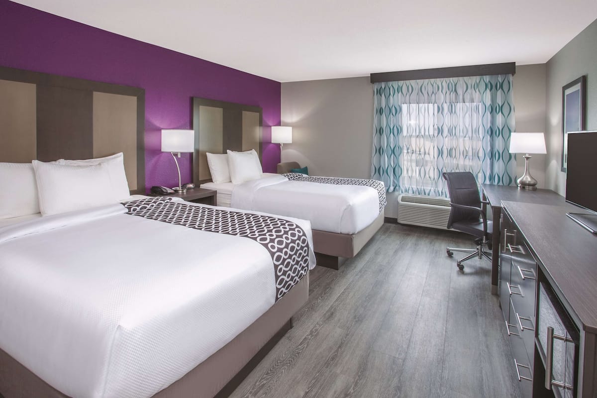 A hotel room with two beds, a desk, a chair, and a large window with patterned curtains.