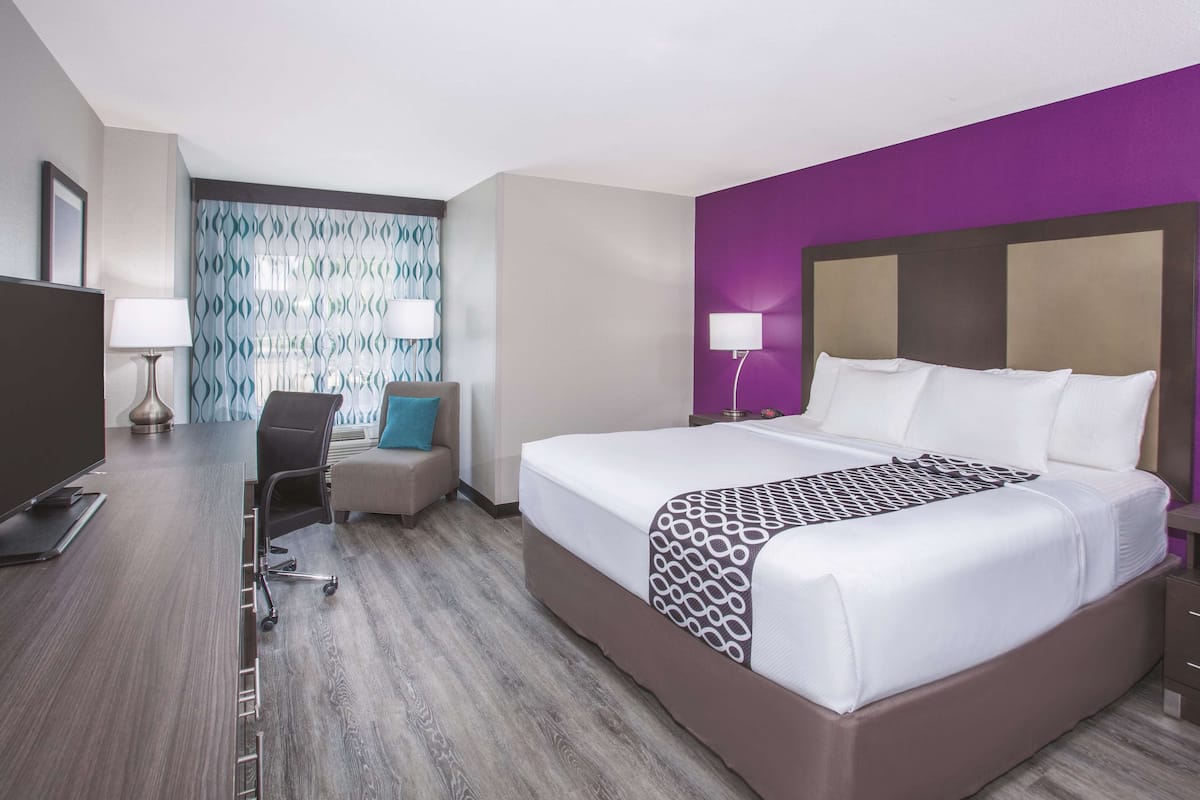A hotel room with a large bed, a desk with a chair, a TV, and a patterned wall.