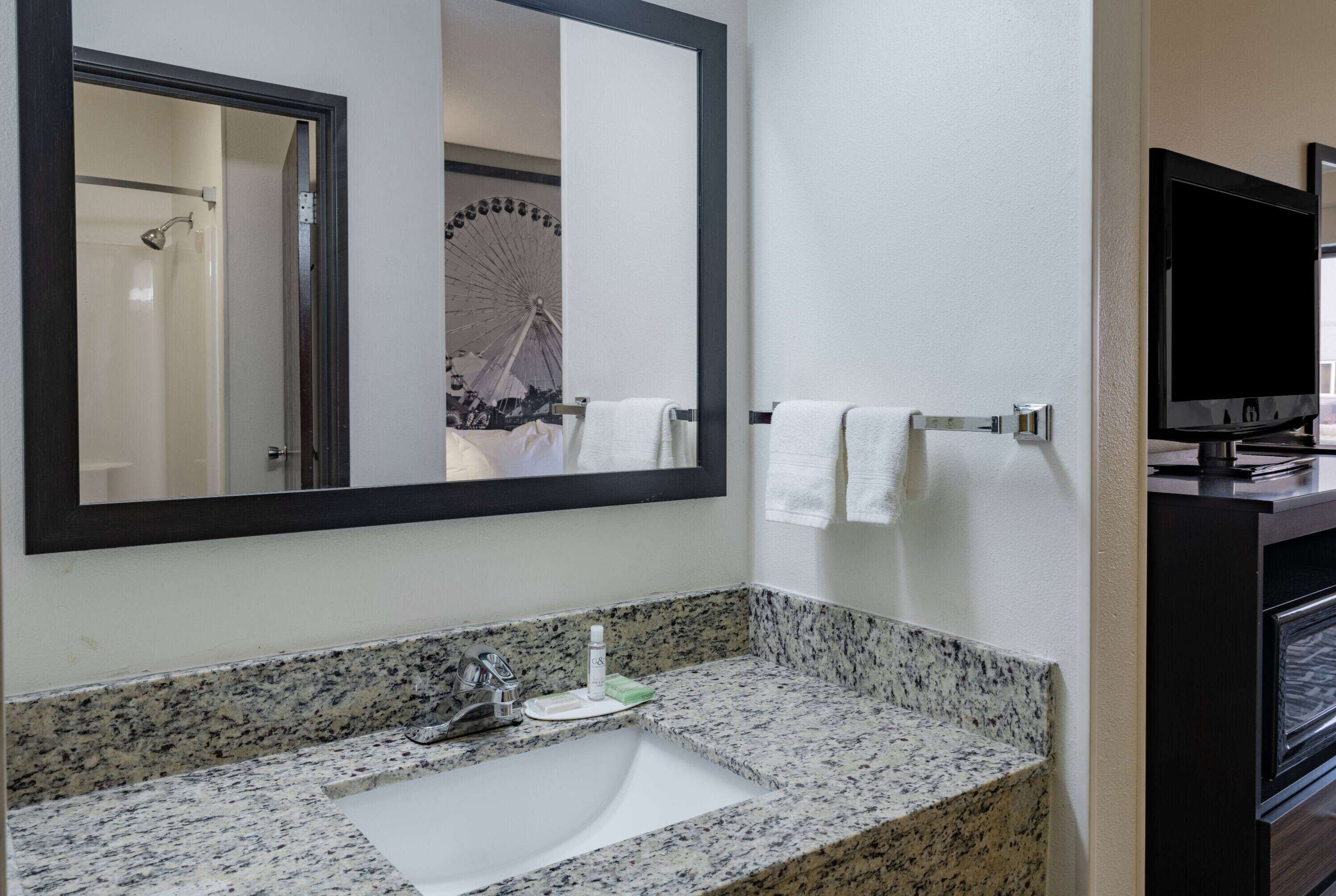 A bathroom with a granite countertop sink, a mirror, a towel rack, and a television.