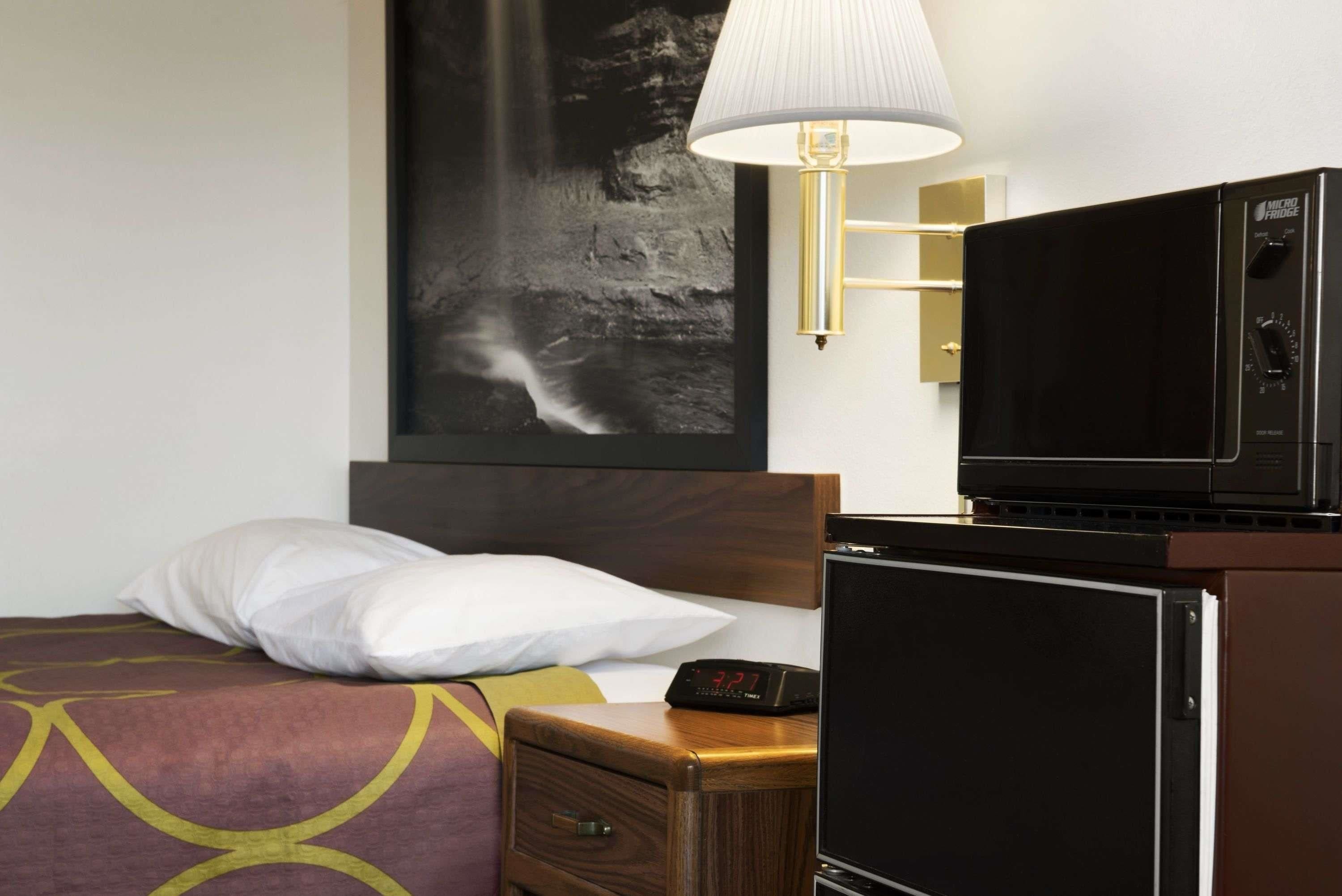 A hotel room with a bed, a nightstand, a TV, a refrigerator, and a wall-mounted lamp.