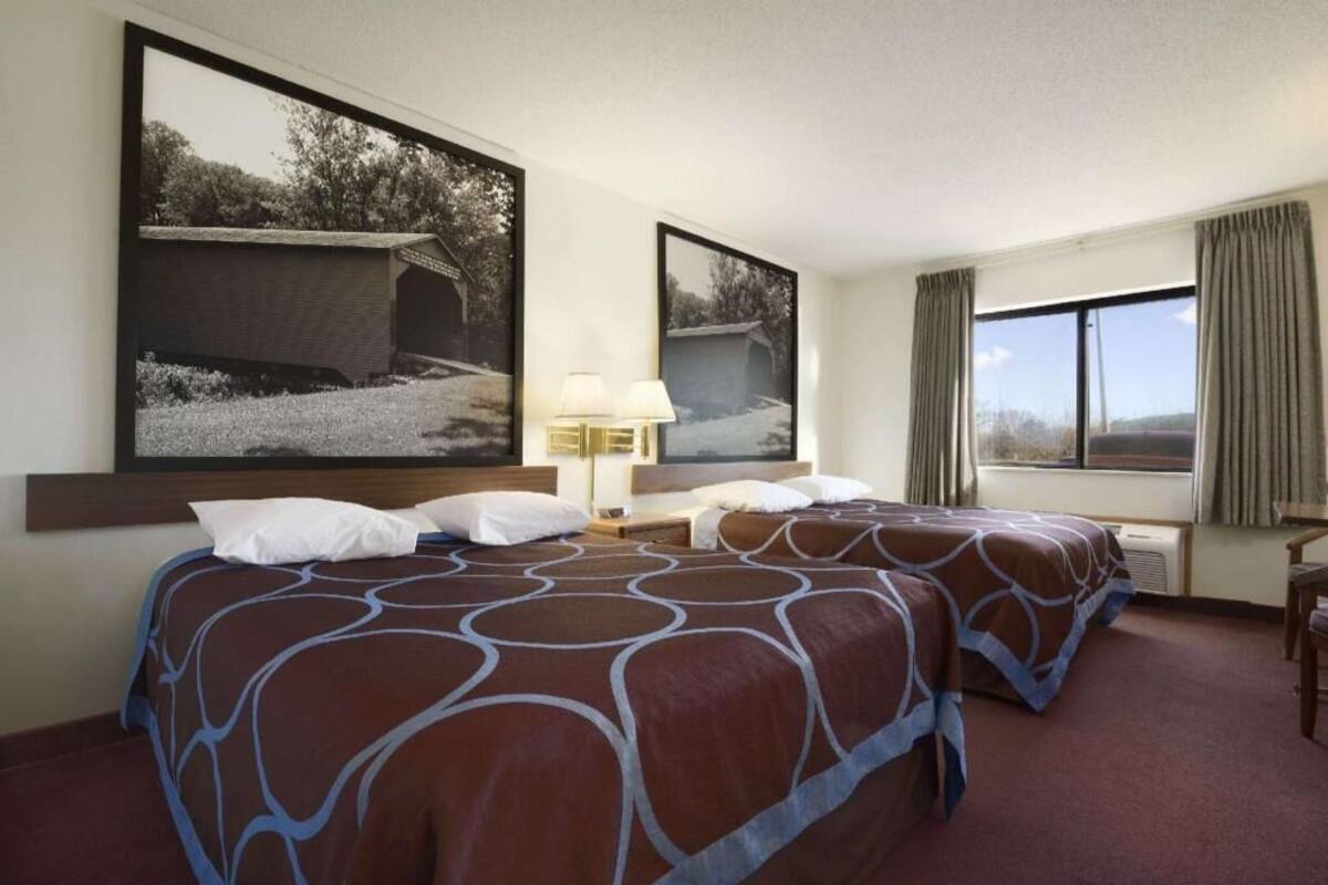 Habitación de hotel con dos camas, un escritorio, una silla, una ventana con cortinas y dos cuadros en la pared.