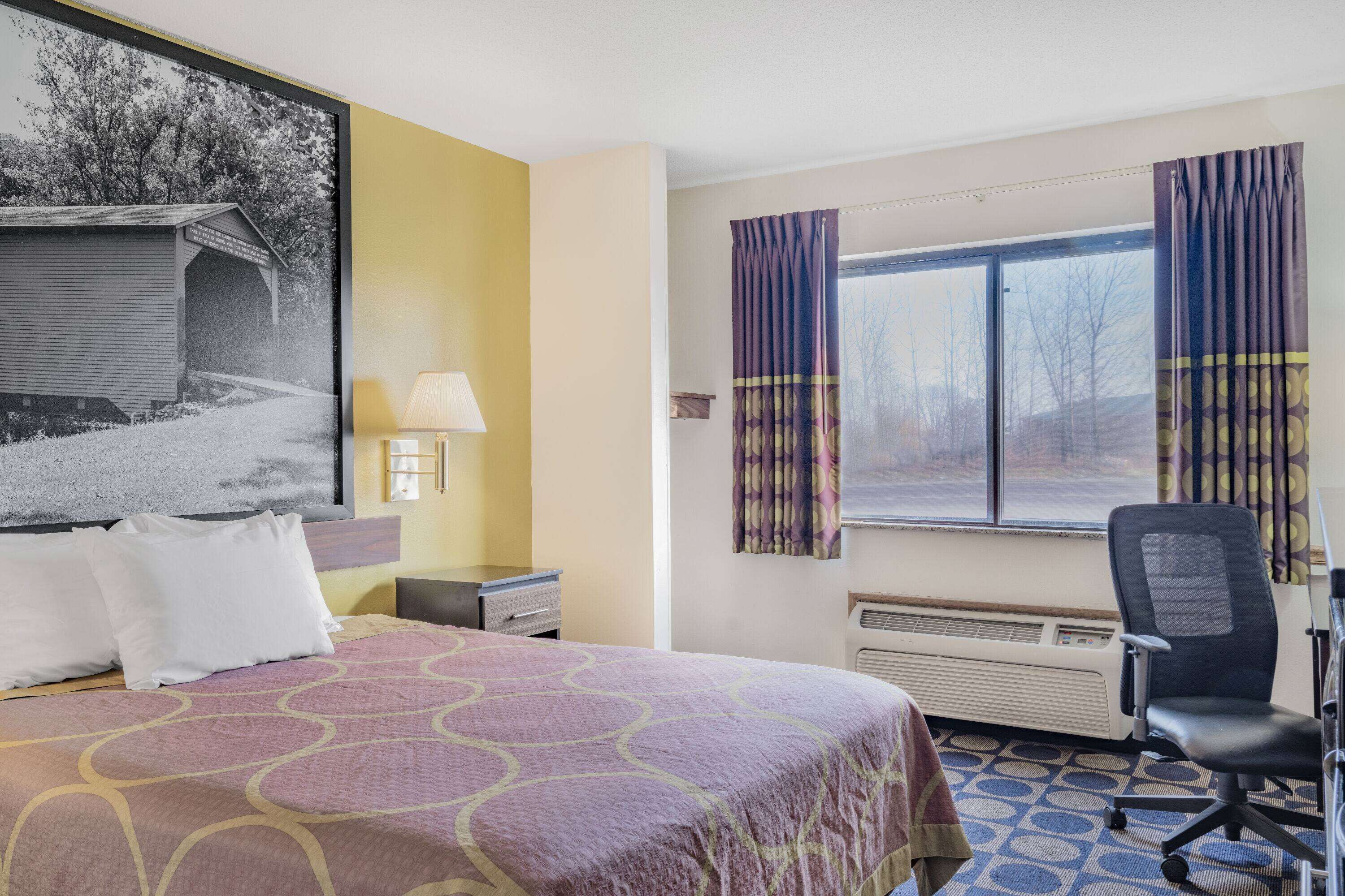A hotel room with a bed, a desk, a chair, a window with curtains, and a wall-mounted lamp.