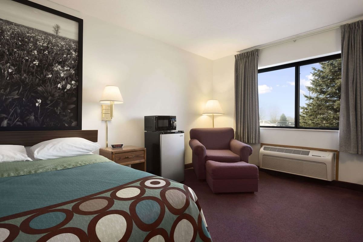 Una habitación de hotel con una cama, una silla, un escritorio, una lámpara, un microondas y un ventanal grande.