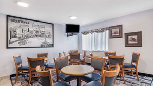 Ein Hotelzimmer mit einem runden Tisch, der von Holzstühlen mit blauen Kissen umgeben ist, einem an der Wand befestigten Flachbildfernseher, einem gerahmten Willkommensschild und einem gemusterten Teppich.