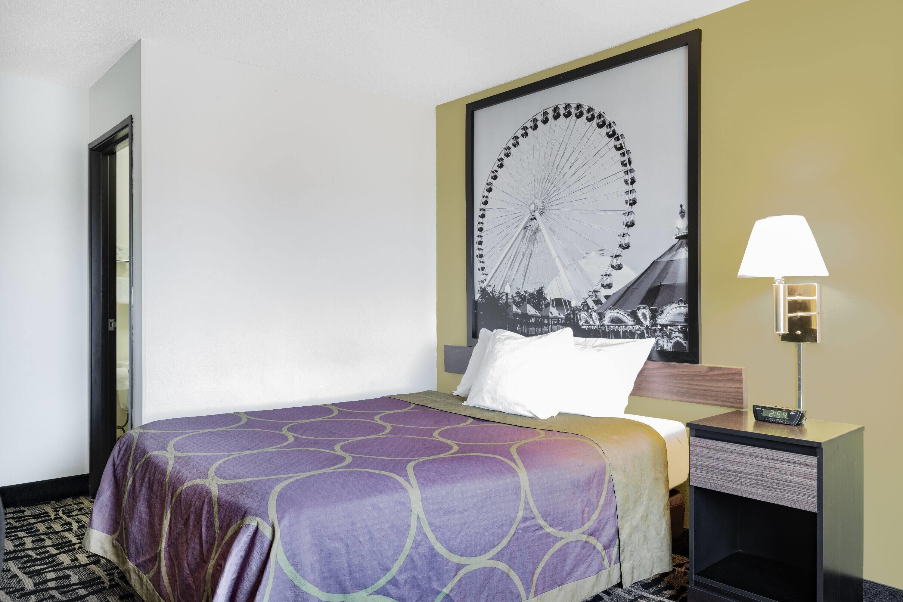 Hotellihuone, jossa on sänky, yöpöytä lampun kanssa, kehystetty kuva näköalapyörästä ja ovi.