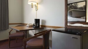 Ein Hotelzimmer mit einem Holztisch, zwei Stühlen, einem Kühlschrank, einer Kaffeemaschine, einer Lampe, einem Telefon und einem Fenster mit Blick auf ein Gebäude und Bäume.