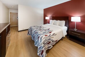 A hotel room with a bed, bedside tables, a nightstand, a wooden dresser, and a door. - Red Roof Inn Fort Wayne (Fort Wayne)