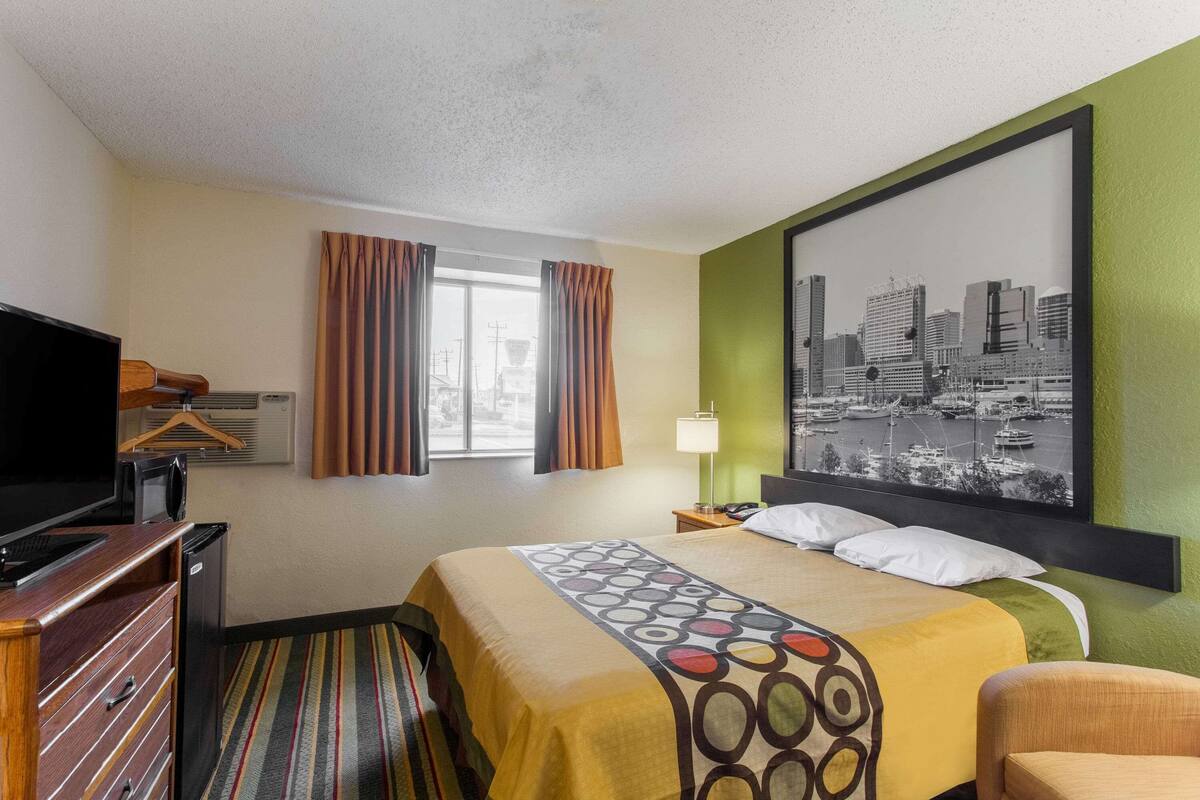 A hotel room with a bed, a television, a desk with a computer, a chair, a window with curtains, and a cityscape painting on the wall.