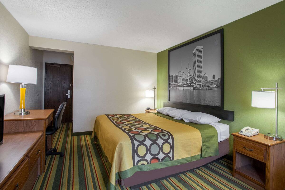 A hotel room with a bed, desk, chair, and a cityscape painting on the wall.