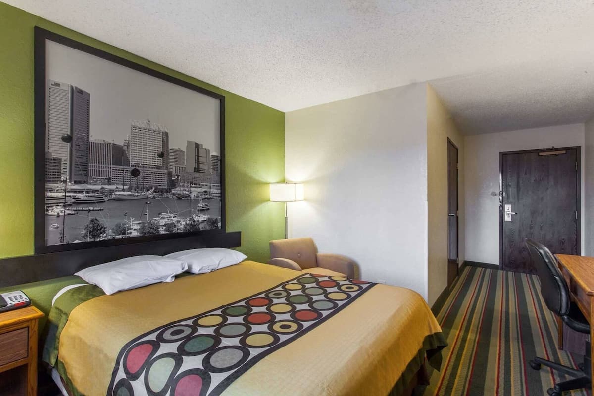A hotel room with a bed, a chair, a desk, a lamp, and a cityscape painting on the wall.