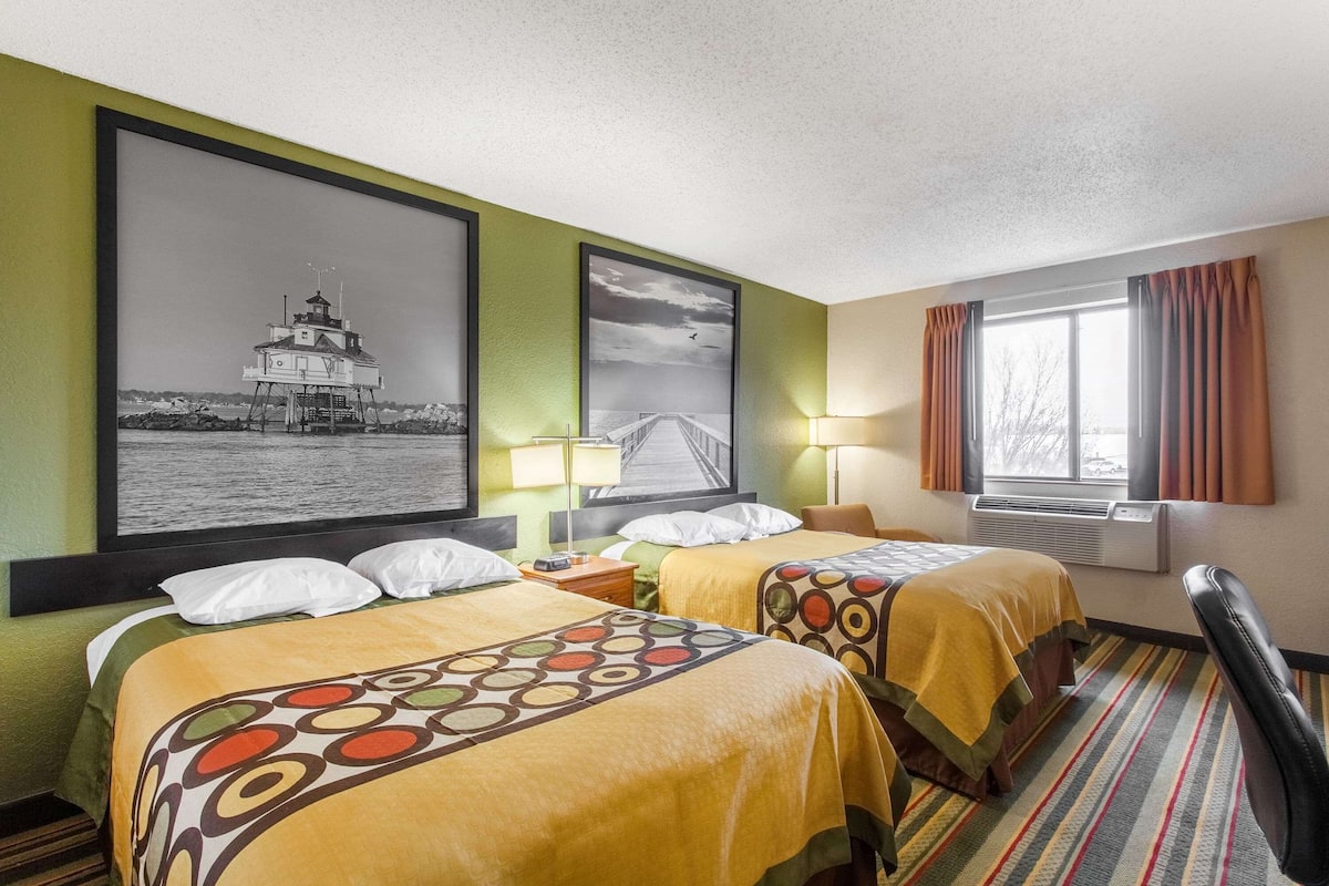 A hotel room with two beds, a large framed picture of a lighthouse, a window with curtains, and a lamp.