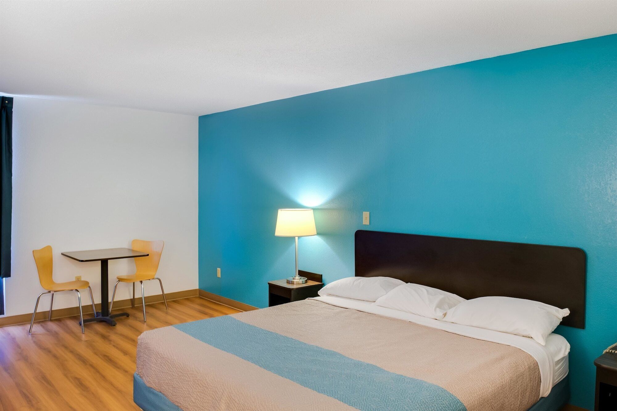 A hotel room with a bed, a desk, two chairs, and a nightstand with a lamp.