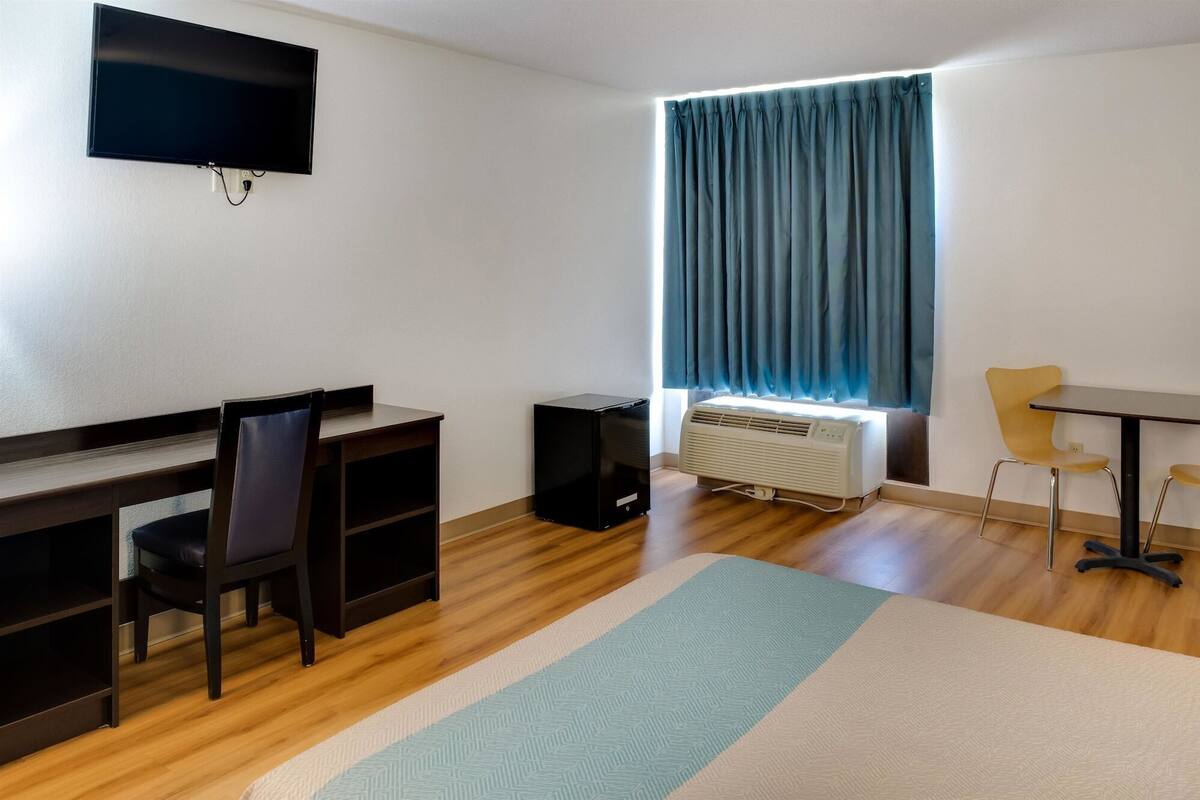 A room with a desk, chair, TV, air conditioning unit, and a window with curtains.