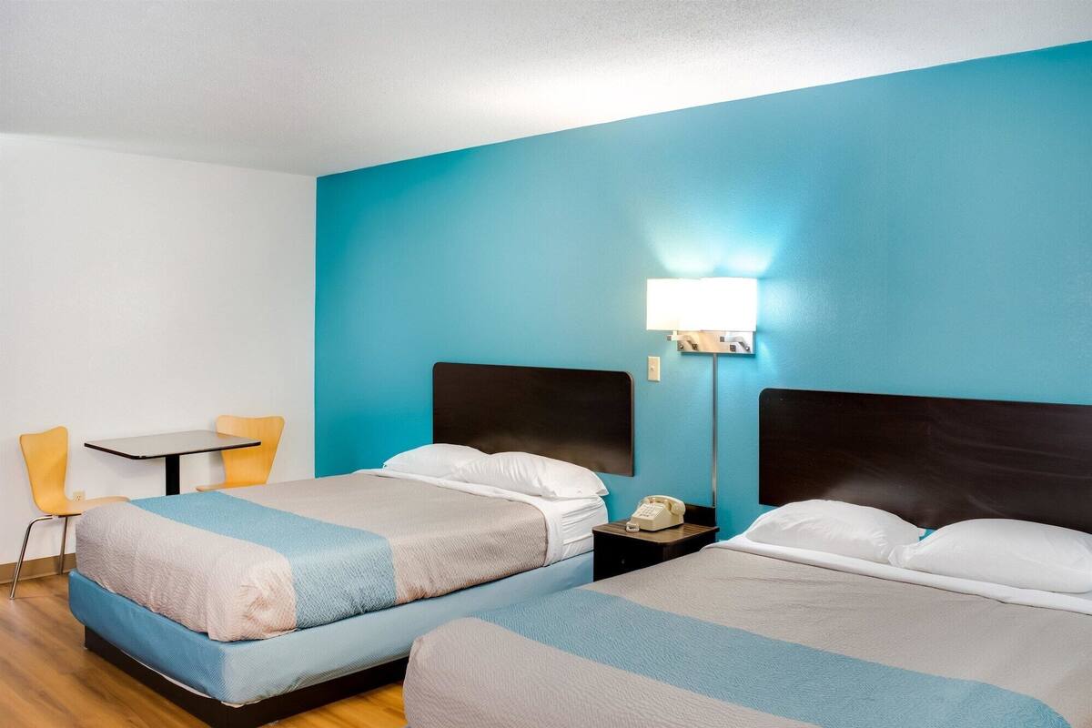 A hotel room with two beds, a desk, a chair, and a nightstand with a lamp.